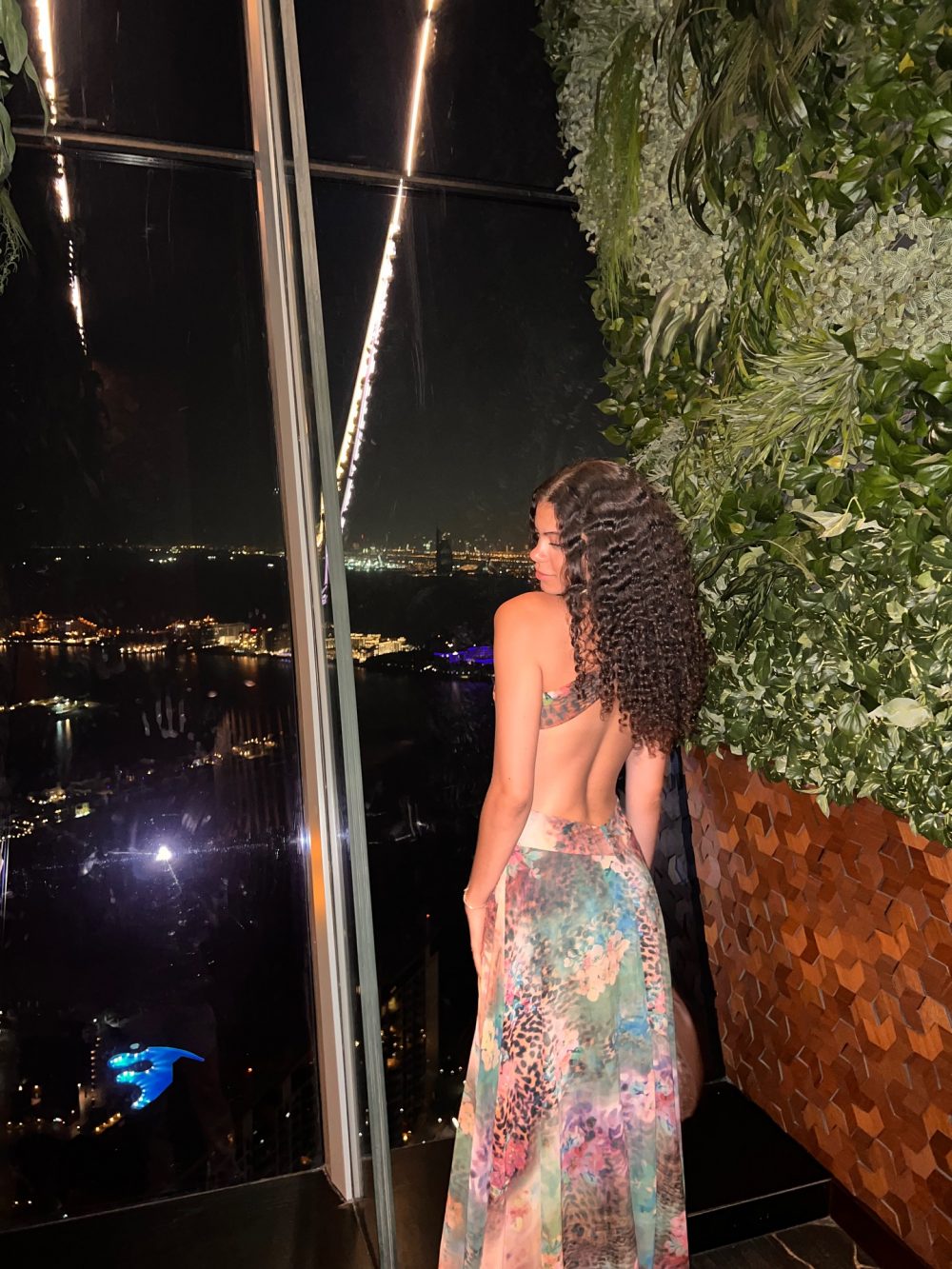 Woman with curly hair in a colorful floral dress stands by a glass window at night, city lights in the background, green wall to the right.