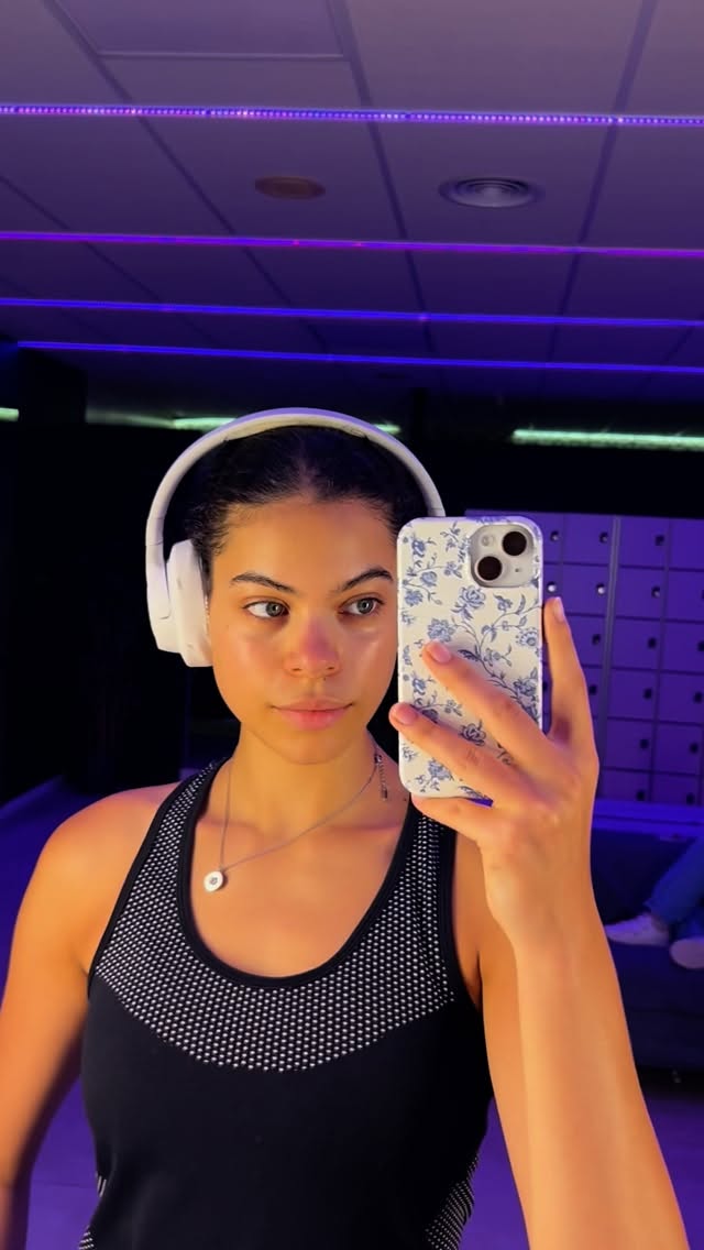 Self-portrait of a woman in a gym taking a mirror selfie with a floral phone case and white over-ear headphones under purple lighting.