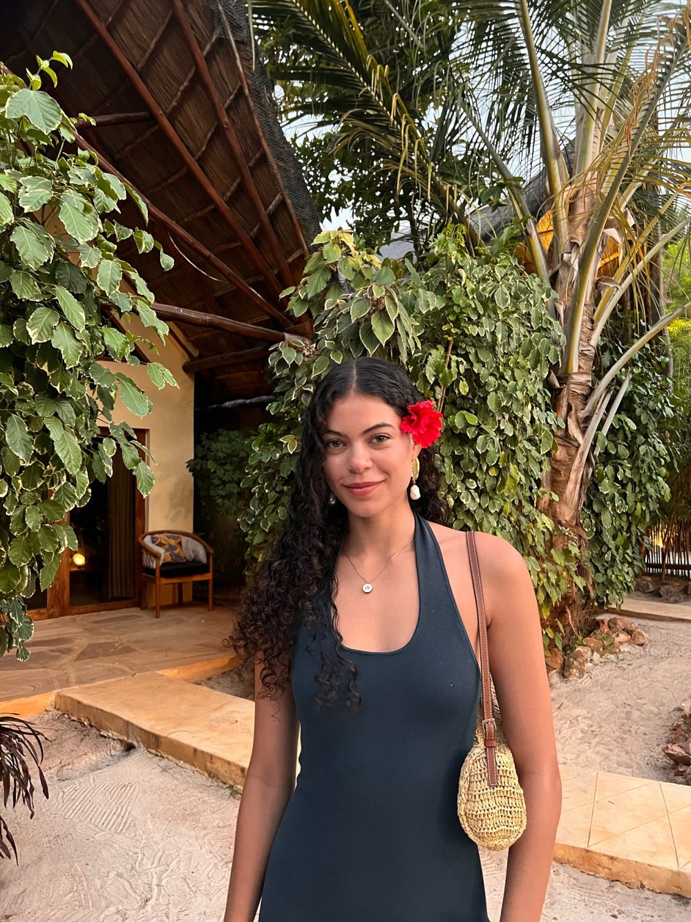 Young woman with long curly hair and a bright red hibiscus in her hair stands among tropical plants outdoors.