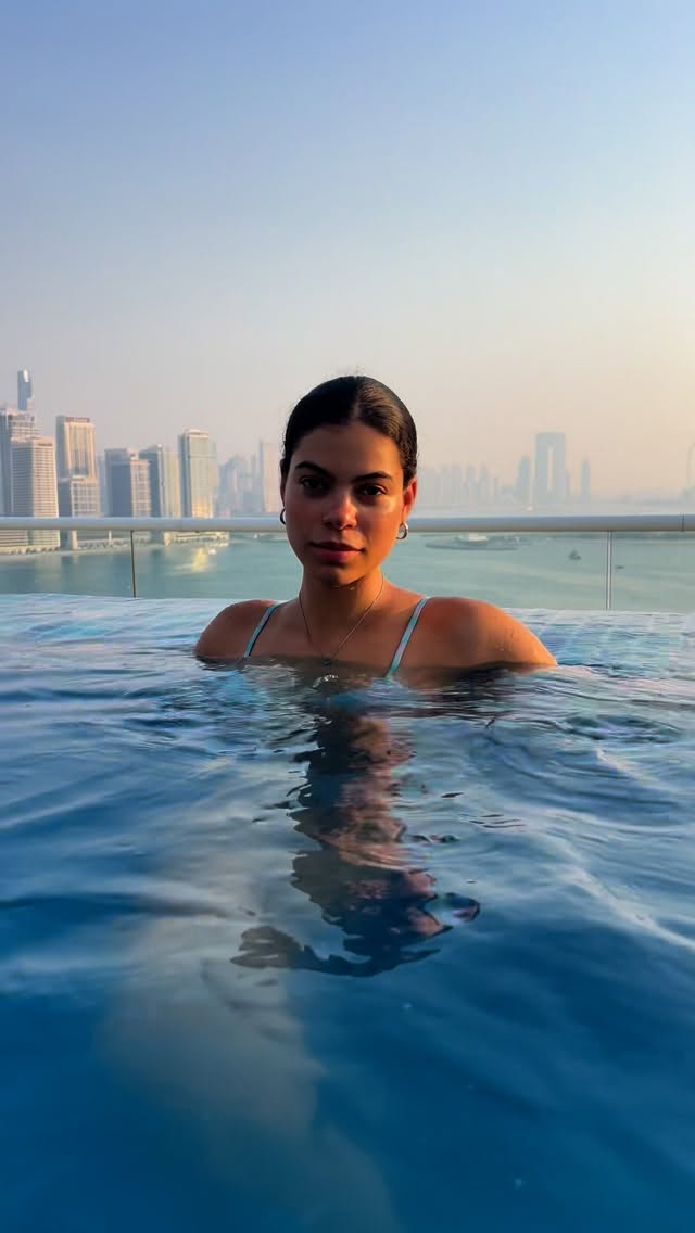 Woman in a blue swimsuit swim top standing in a rooftop pool, city skyline and river in the background at dusk/light haze.