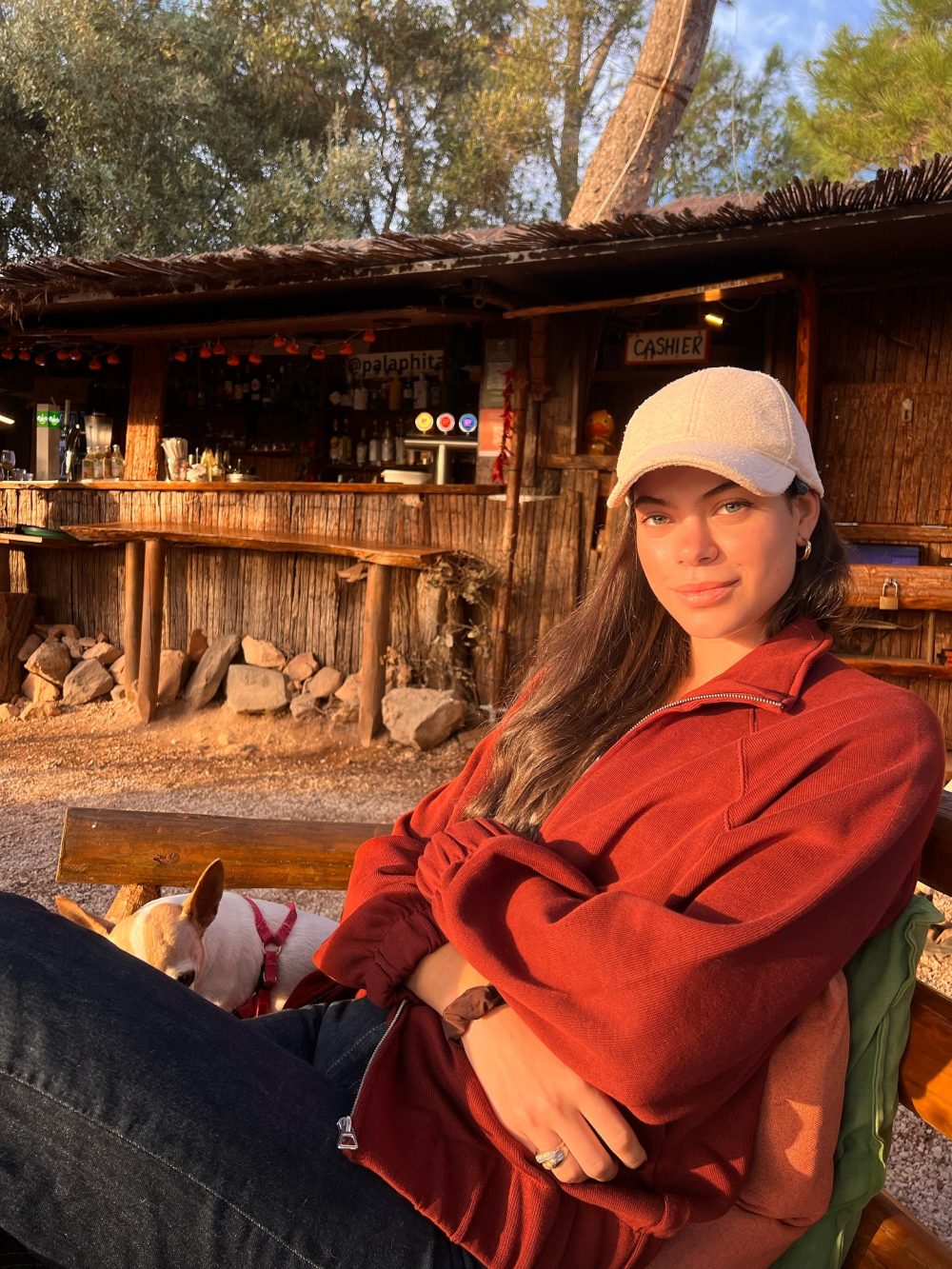 Woman in red jacket sits on a wooden bench at an outdoor rustic bar, with a small dog nearby; warm sunlight on her face.