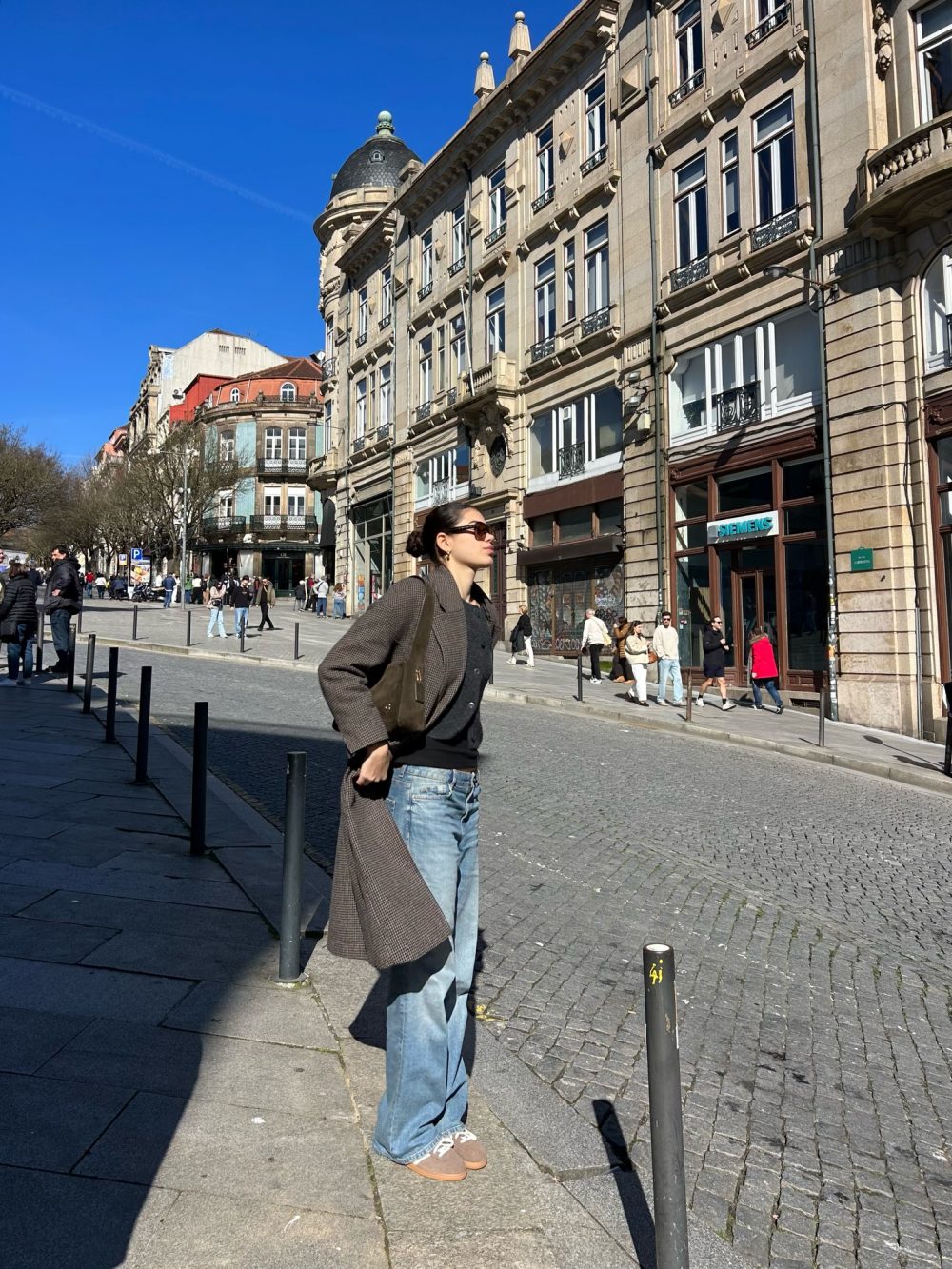 Young woman wearing a brown coat and sunglasses walks along a sunny European street with ornate stone buildings.