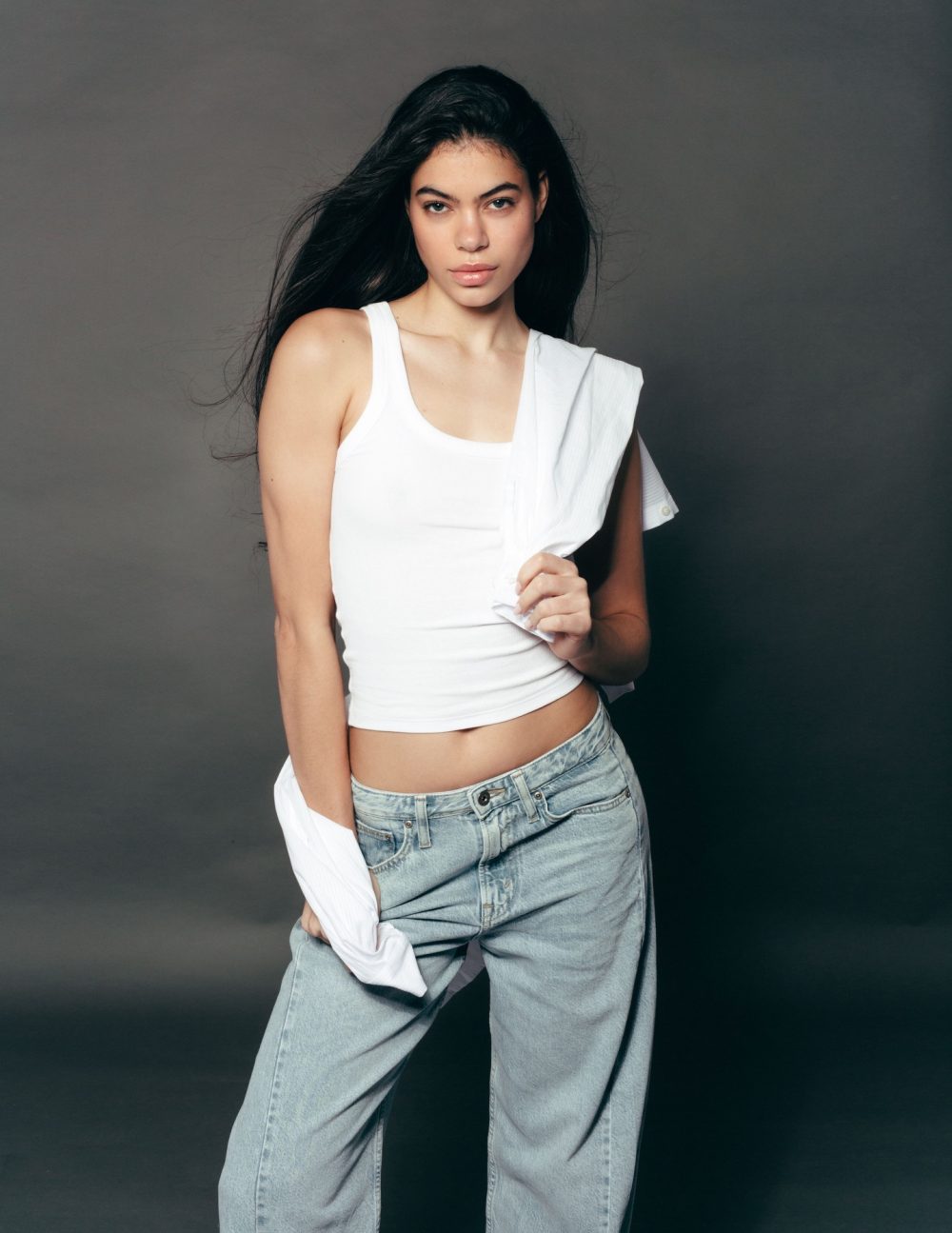 Young woman with long dark hair in a white tank top and jeans, holding a white shirt over her shoulder against a gray backdrop.