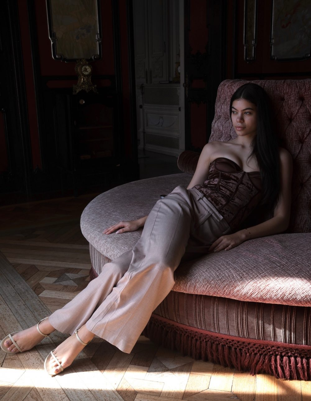 Young woman reclining on a pink tufted sofa in a dimly lit, ornate room.