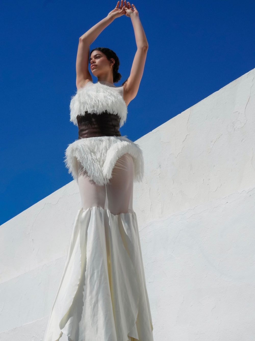 Woman in a white fluffy dress with a dark waist belt raises arms against a bright blue sky and white wall.