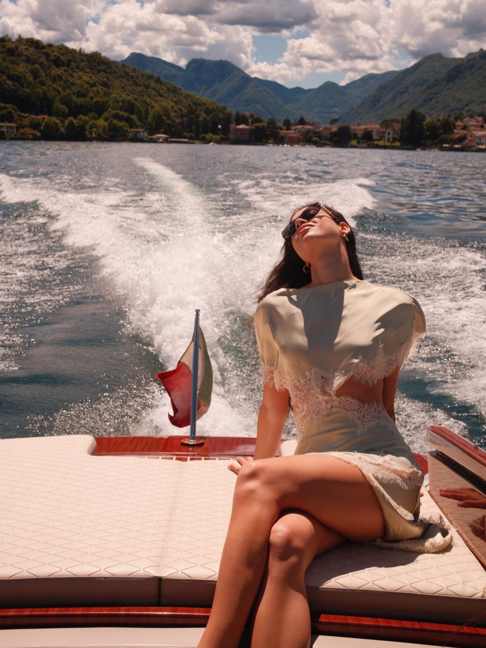 Woman relaxing on a boat seat, head tilted back, wearing sunglasses and a beige dress by the lake with misty mountains in the background.