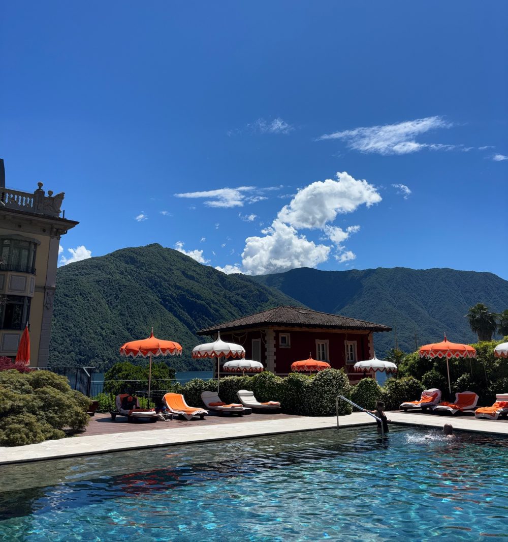 Resort pool area with orange and white umbrellas, lounge chairs, and a mountain backdrop under a bright blue sky