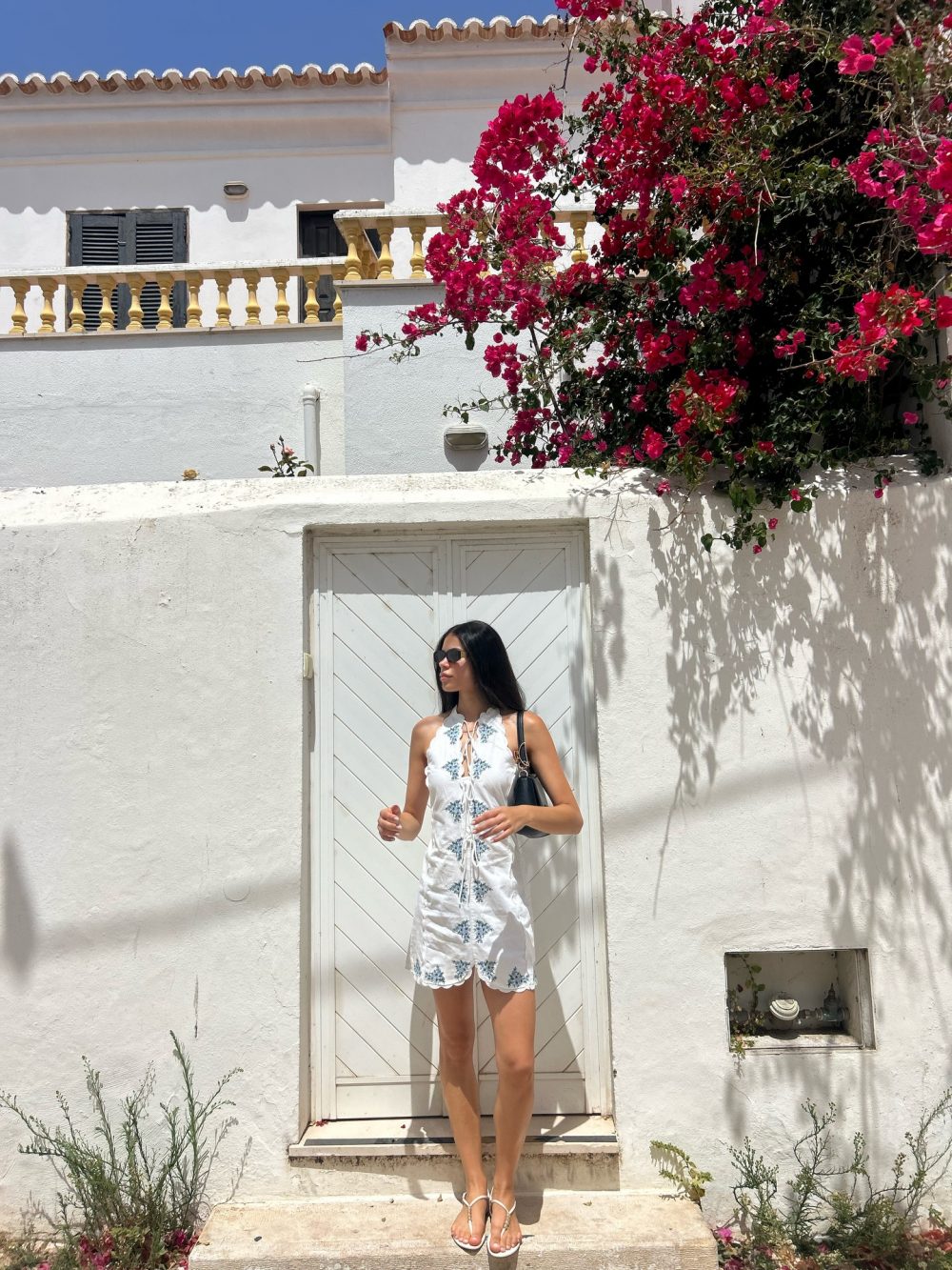 Woman in a white embroidered sundress and sandals stands on a stone step in front of a white wall with a door, pink bougainvillea spilling over the right side.