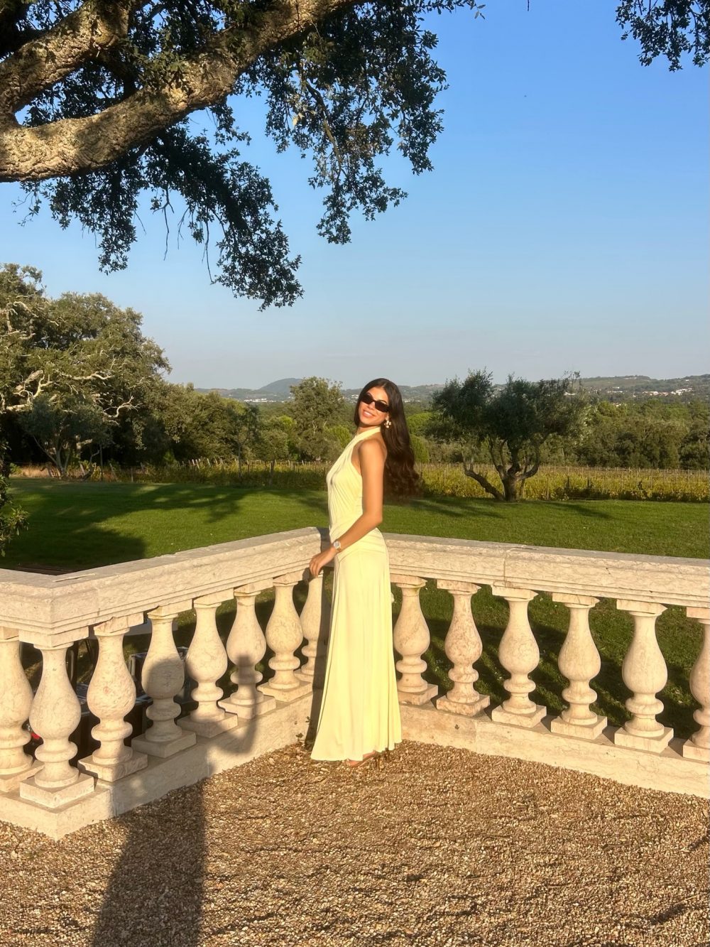 Woman in a pale yellow dress stands by a stone balustrade, with a sunny garden and distant hills in the background
