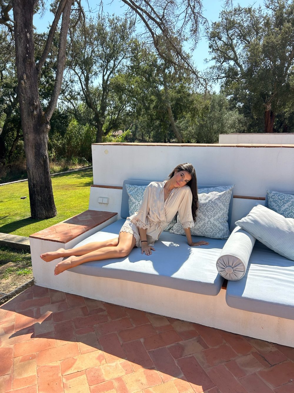 Woman in a cream robe reclining on a light blue outdoor sofa on a sunny patio with trees behind the scene in the background.