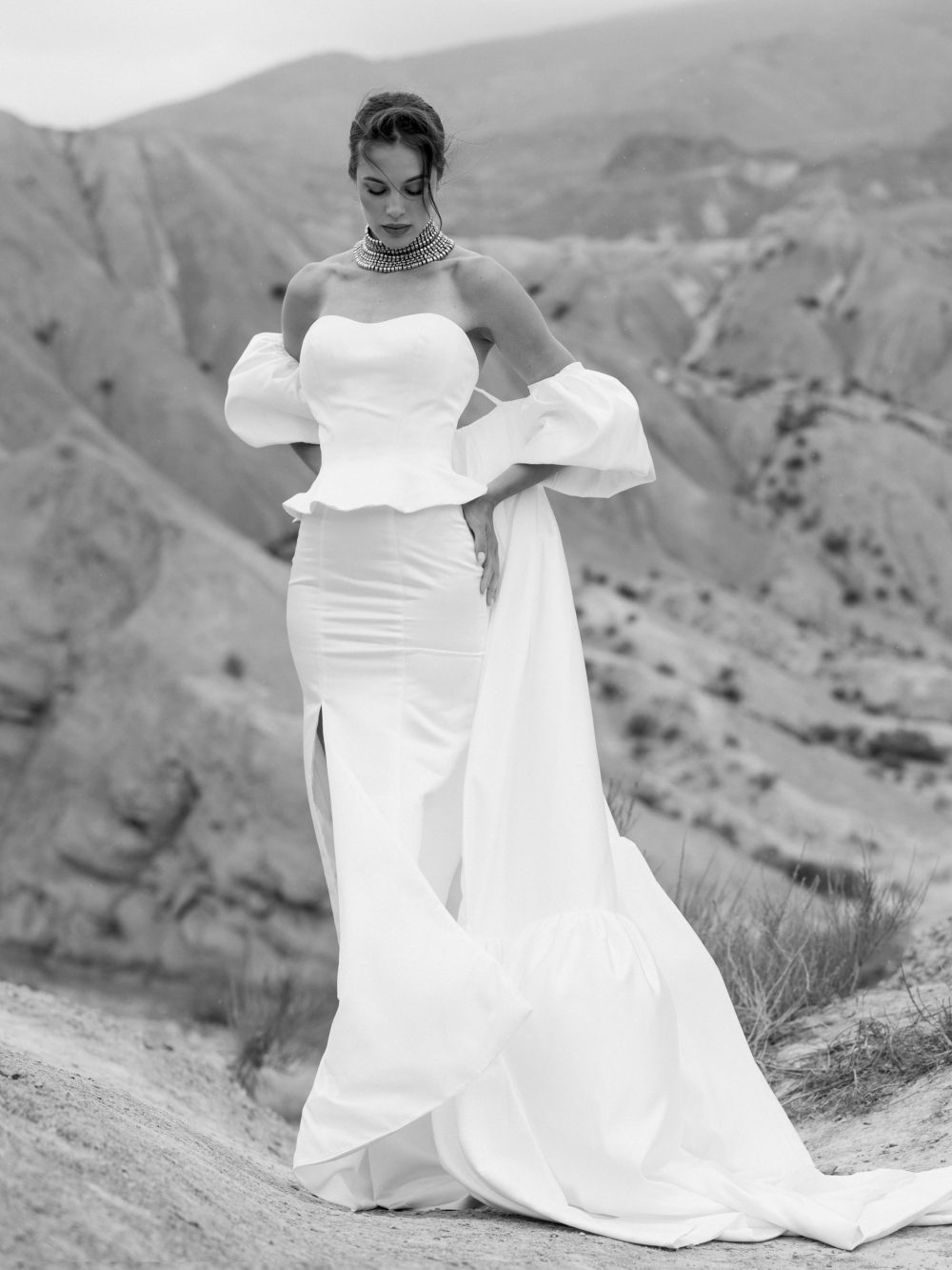 Bride in a strapless white wedding gown with dramatic puff sleeves, standing on a rocky desert hillside.