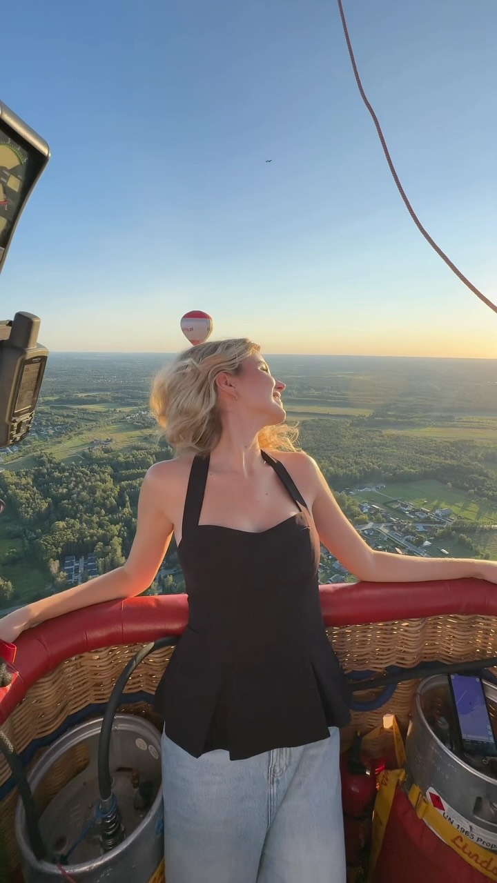 Woman in a black top and light jeans leaning on a red wicker hot air balloon basket rim, sunset over a green landscape.
