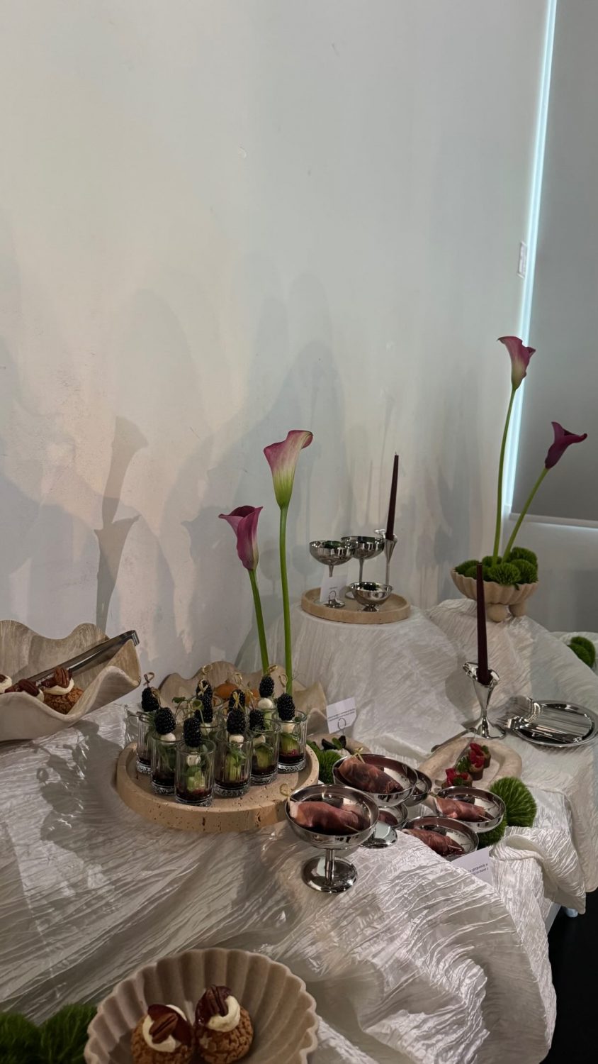 Buffet table with a white textured tablecloth, tall pink calla lilies, and small dessert glasses arranged on rounds around the centerpiece.