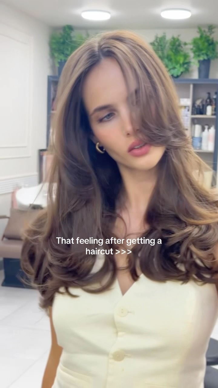 Woman with long brown wavy hair in a hair salon, wearing a light yellow button-up top, looking slightly to the side as if showing off a fresh haircut.