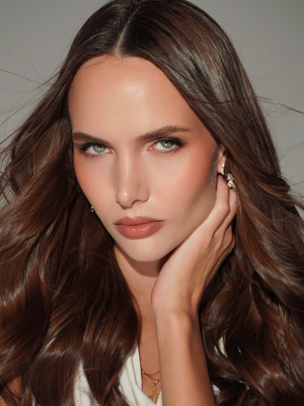 Close-up of a woman with long brown hair, green eyes, and subtle makeup, resting her face on her hand and wearing a gold cross necklace and diamond earrings.