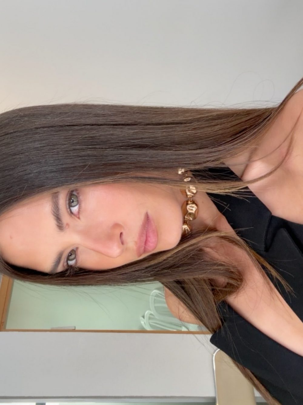 Close-up portrait of a woman with long brown hair wearing a gold chunky necklace and a black top, looking at the camera indoors.