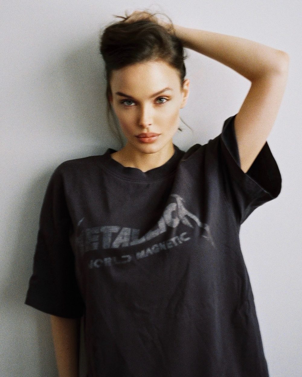 Portrait of a woman with dark hair in a loose, dark graphic T-shirt, one arm raised behind her head against a gray wall.