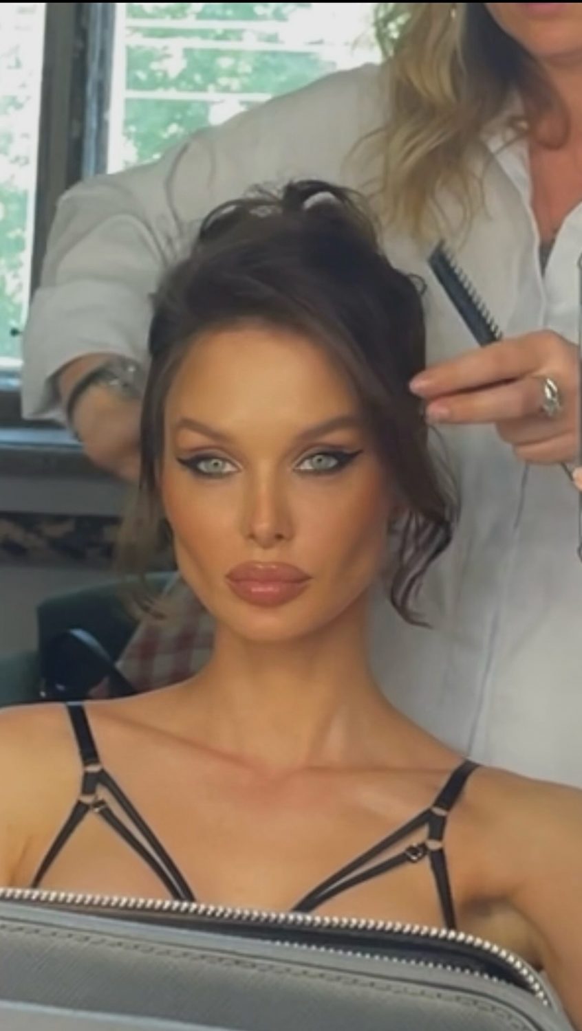 Close-up of a woman with dramatic makeup and blue eyes in a salon, hair being styled by a stylist holding a comb behind her head.