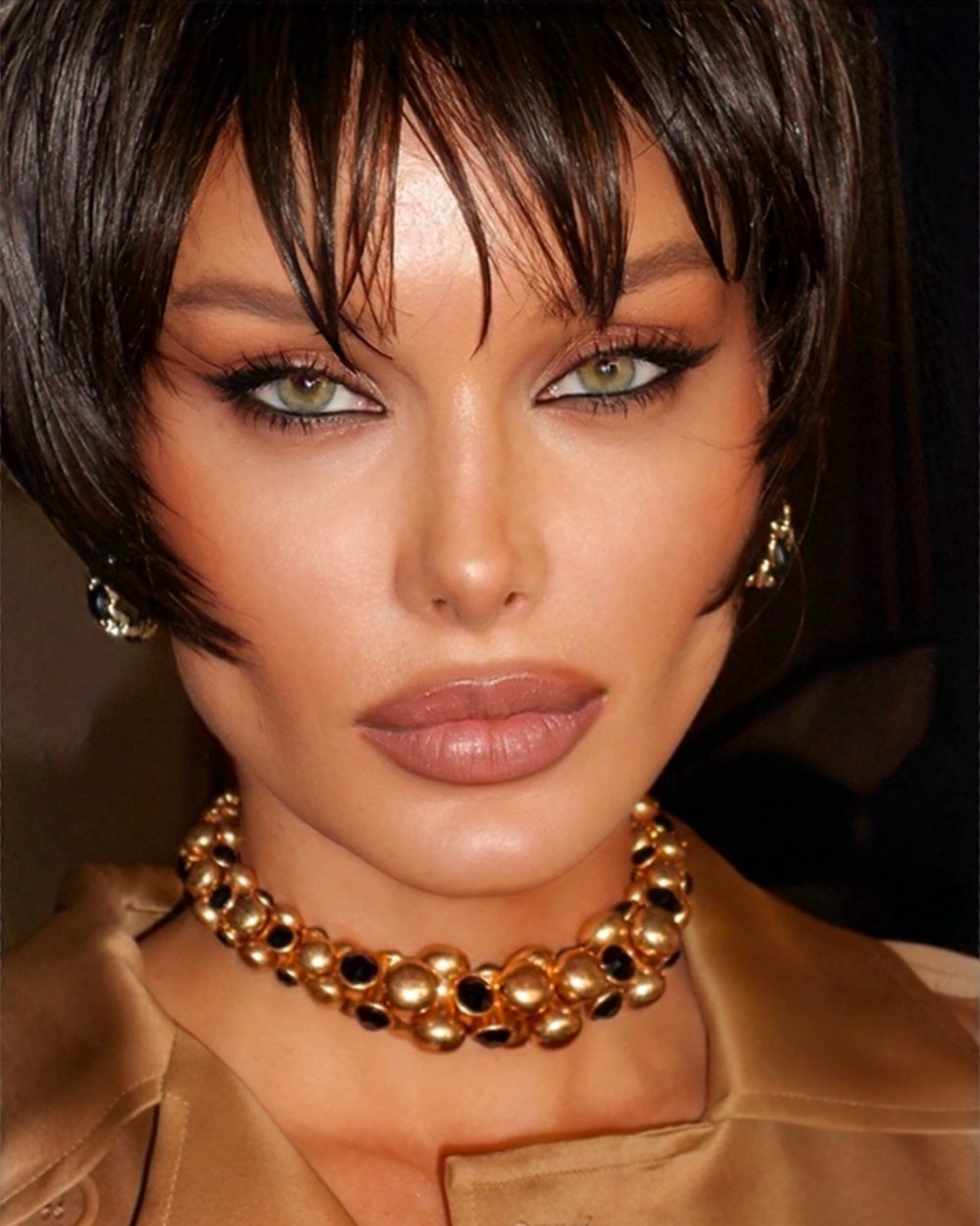 Close-up of a woman's face focusing on green eyes with dark eyeliner, wearing a gold necklace and beige satin top.