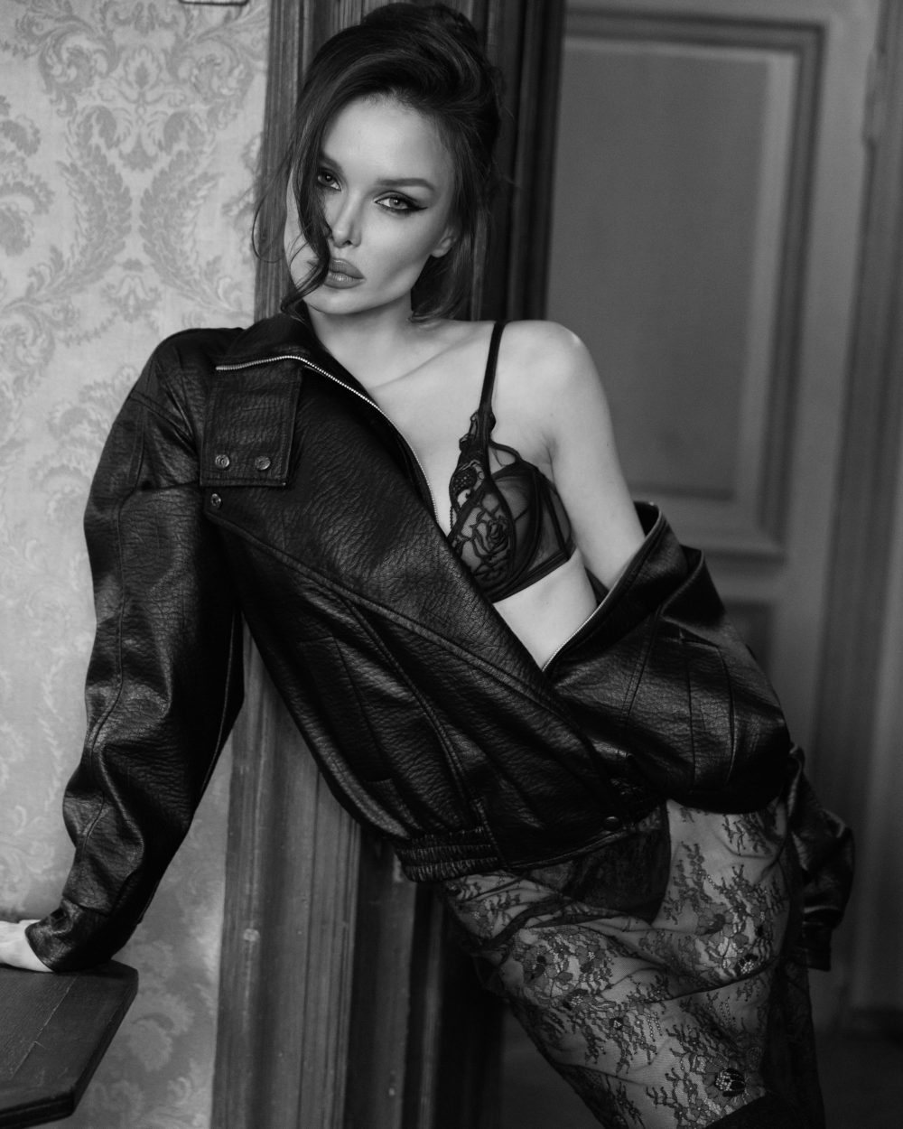 Black-and-white photo of a woman in lace lingerie and a leather jacket posing against a wooden door.