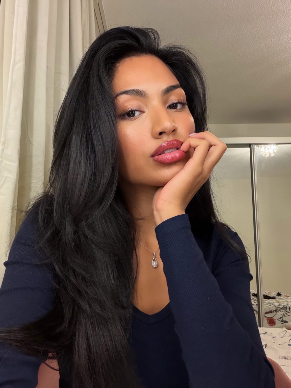 Portrait of a woman with long black hair, navy sweater, pink lipstick, resting her chin on her hand in a bathroom setting.