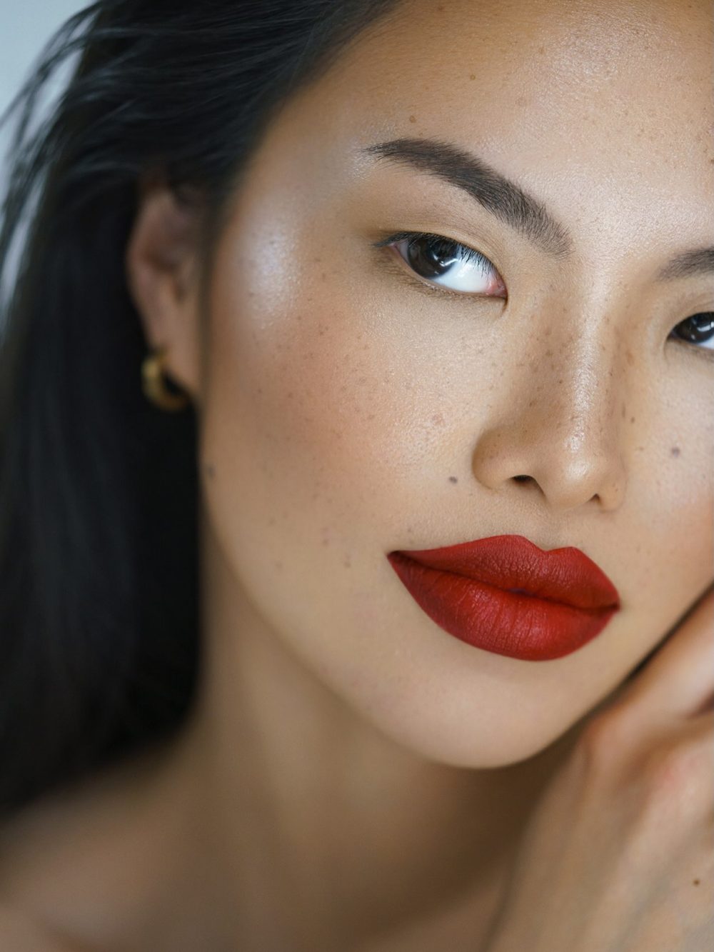 Close-up of a woman with bold red lipstick, freckles, and dark hair, looking slightly to the left.