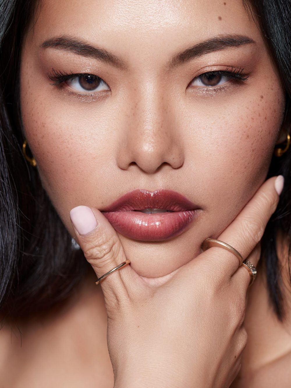 Close-up portrait of a woman with warm makeup, freckles, and a hand resting on her chin, highlighting lips and eyes.