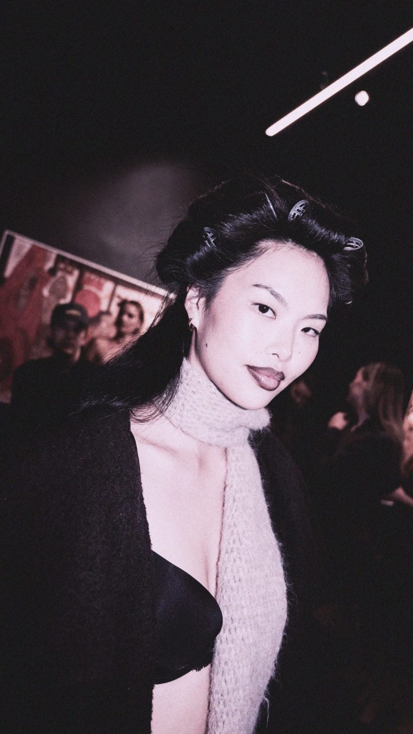Portrait of a woman with dark hair in rollers, wearing a cream scarf and black top in a dim backstage setting.