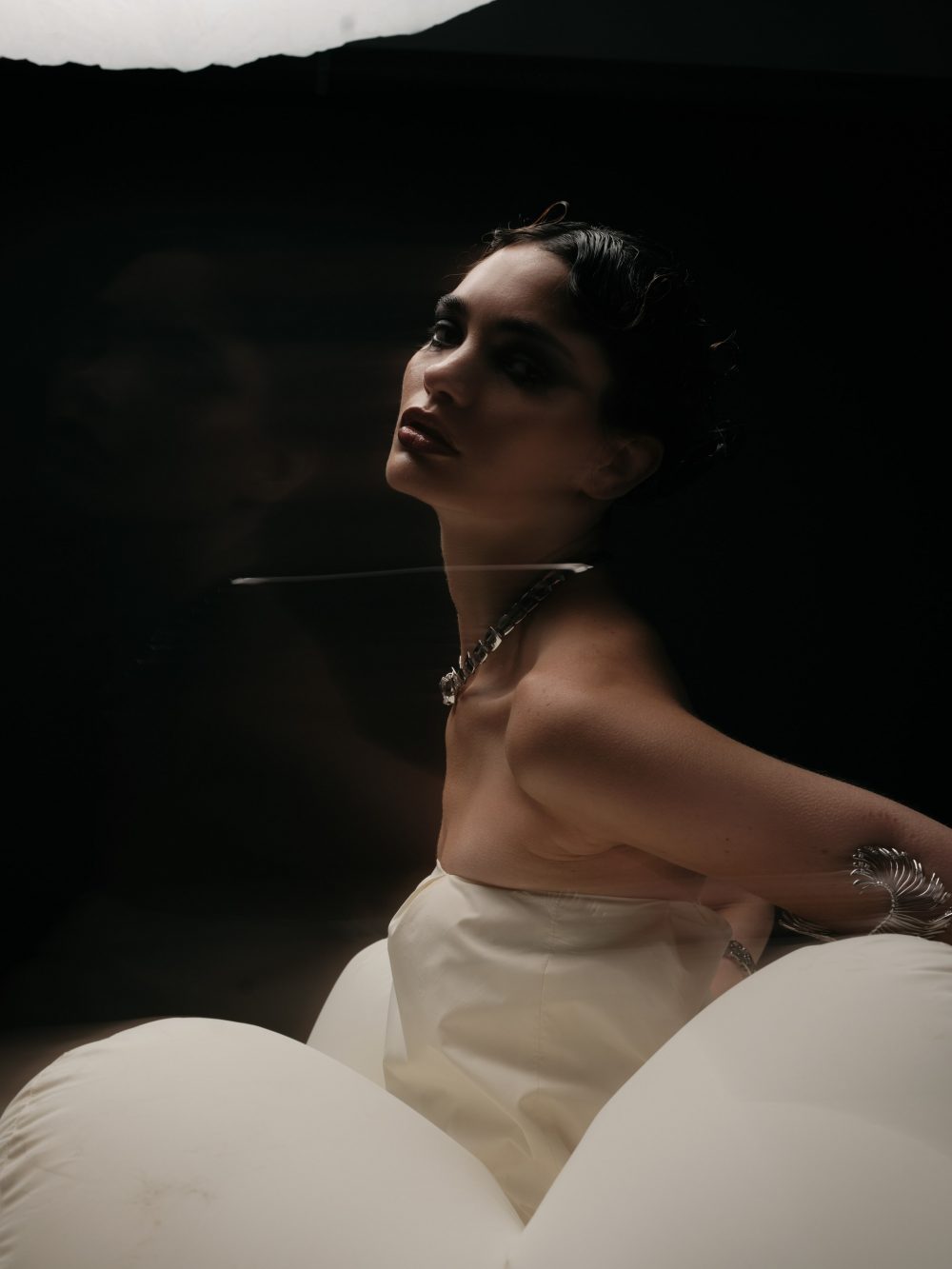 Elegant woman in a white gown with a silver necklace tilts her head back against a dark backdrop, dramatic lighting. (informative fashion portrait)