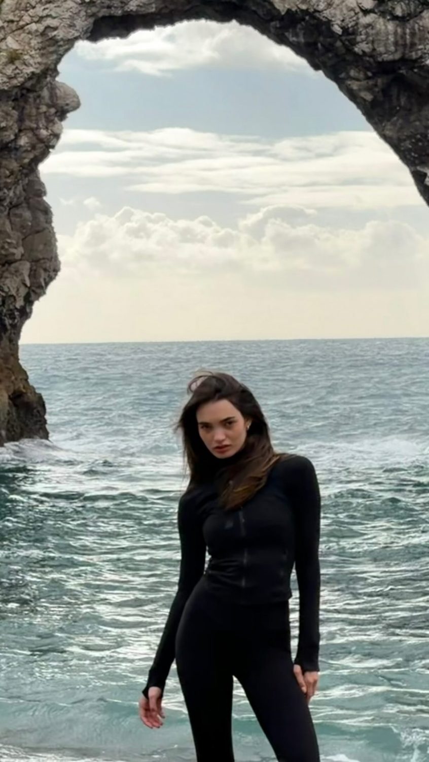 Person in a black wetsuit standing in shallow water near a rocky sea arch with a cloudy sky in the background.