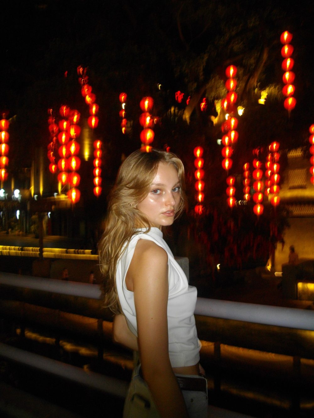 Woman with blonde hair in a white sleeveless top posing at night with red lanterns in the background.