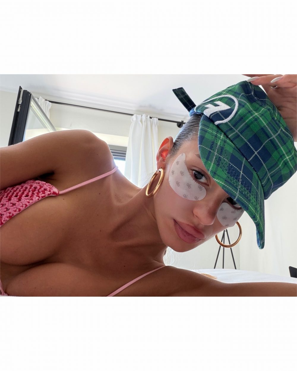 Two people taking a playful selfie close up, wearing a green plaid cap, pink bikini top, and gold hoop earrings in a bright room