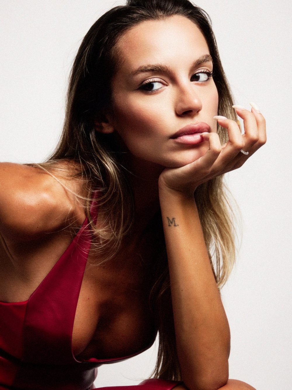Close-up portrait of a woman with long hair resting her chin on her hand, wearing a red dress, tattooed 'M' on her forearm visible.