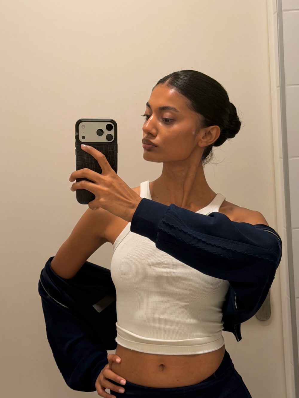 Woman in a white tank top taking a mirror selfie in a bathroom, hair slicked back in a bun, wearing a dark jacket draped over shoulders.
