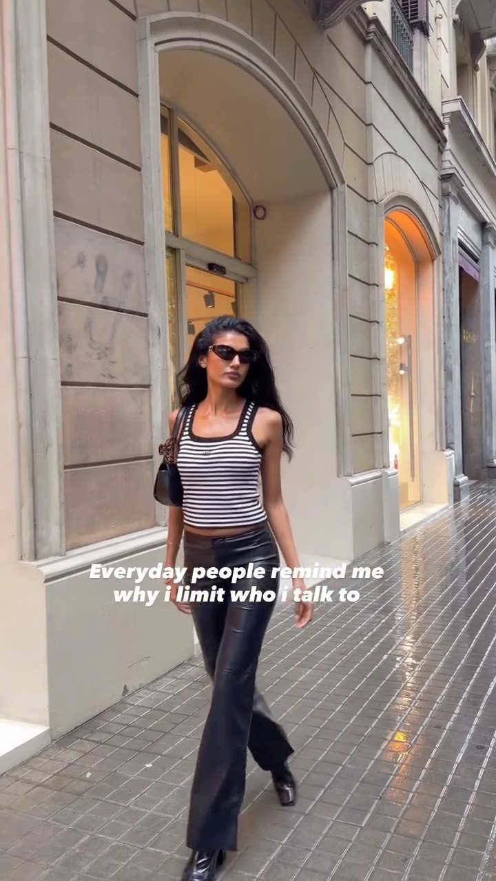 Woman in a striped crop top and black leather pants walks down a city sidewalk wearing sunglasses and a small black purse.
