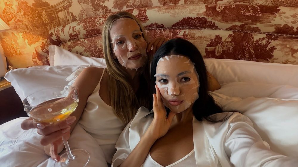 Two women in white robes wear facial sheet masks while posing on a bed; one holds a wine glass toward the camera.