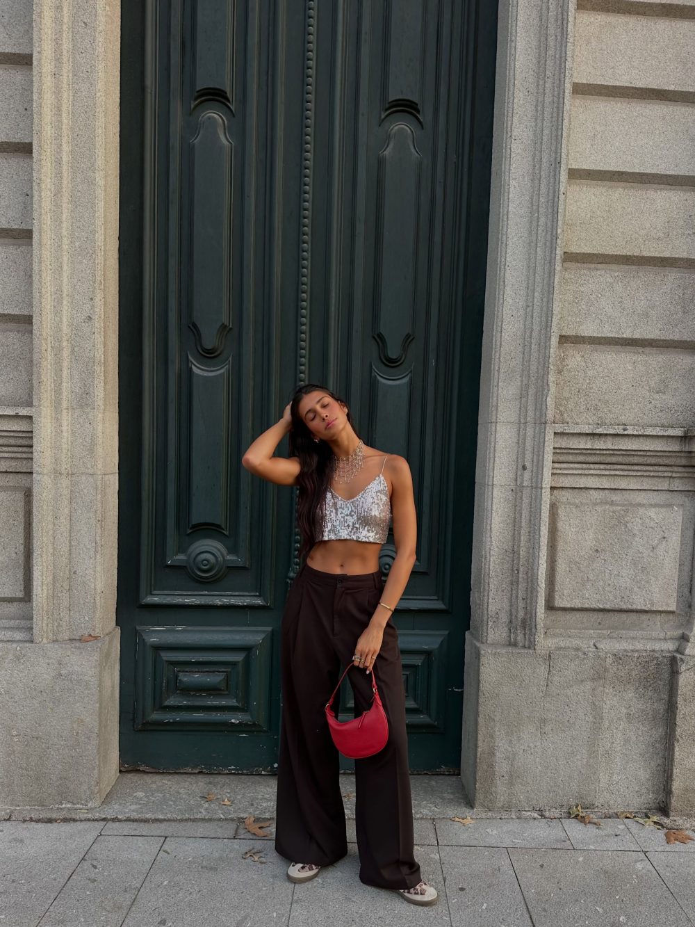 Woman in a silver sequined crop top and wide-leg pants posing against a dark ornate door, holding a red handbag on a city street.