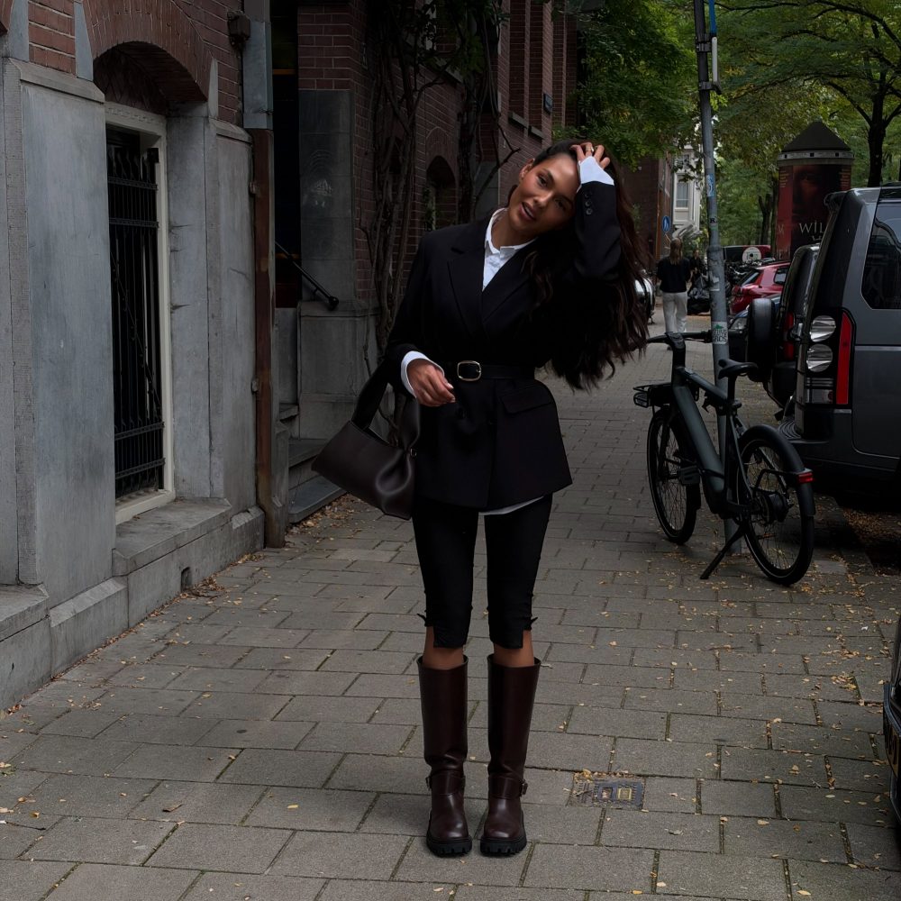 Woman in a black coat and white shirt posing on a city sidewalk with brick buildings and bikes nearby.