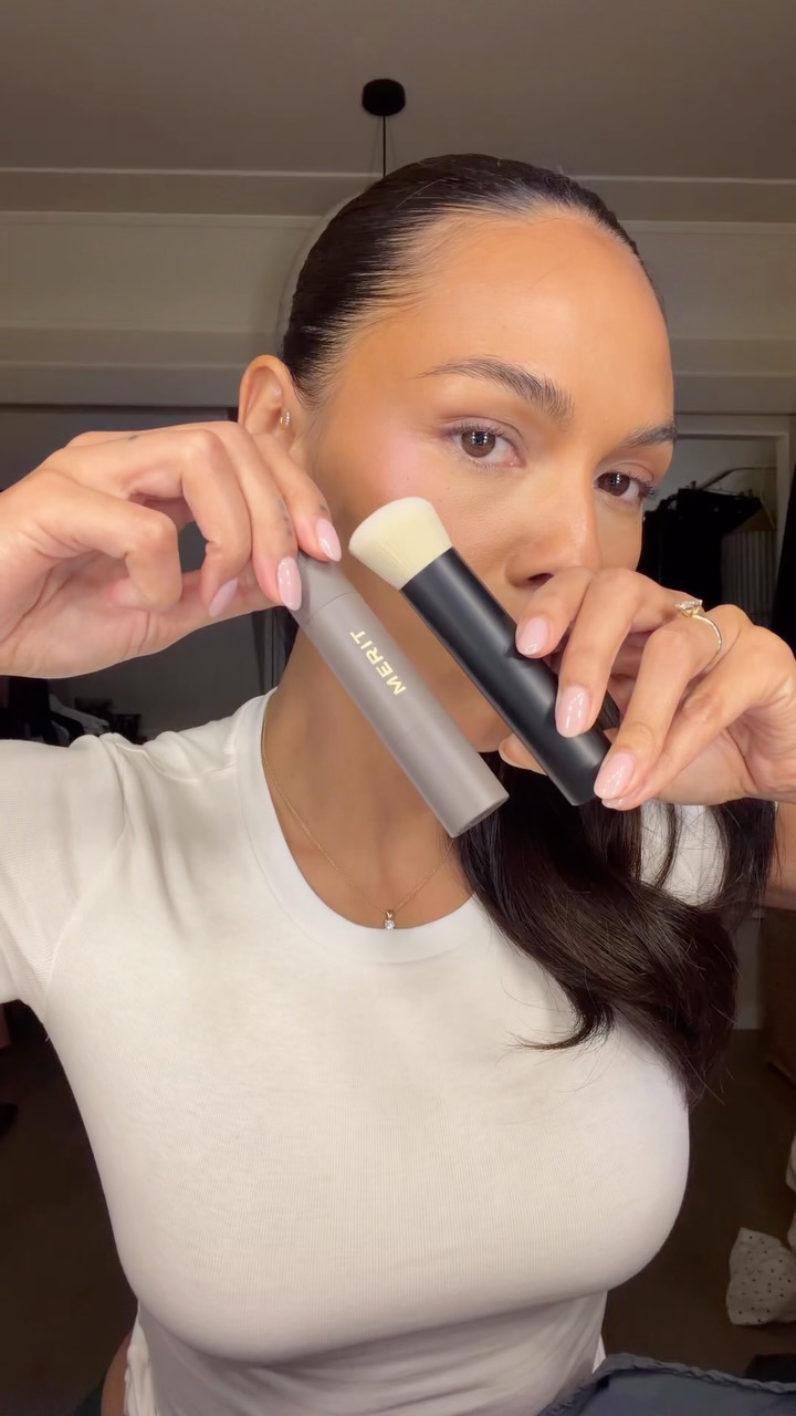 Close-up of a woman with slicked-back hair applying makeup with two brushes; a MERIT product is visible in her hand.