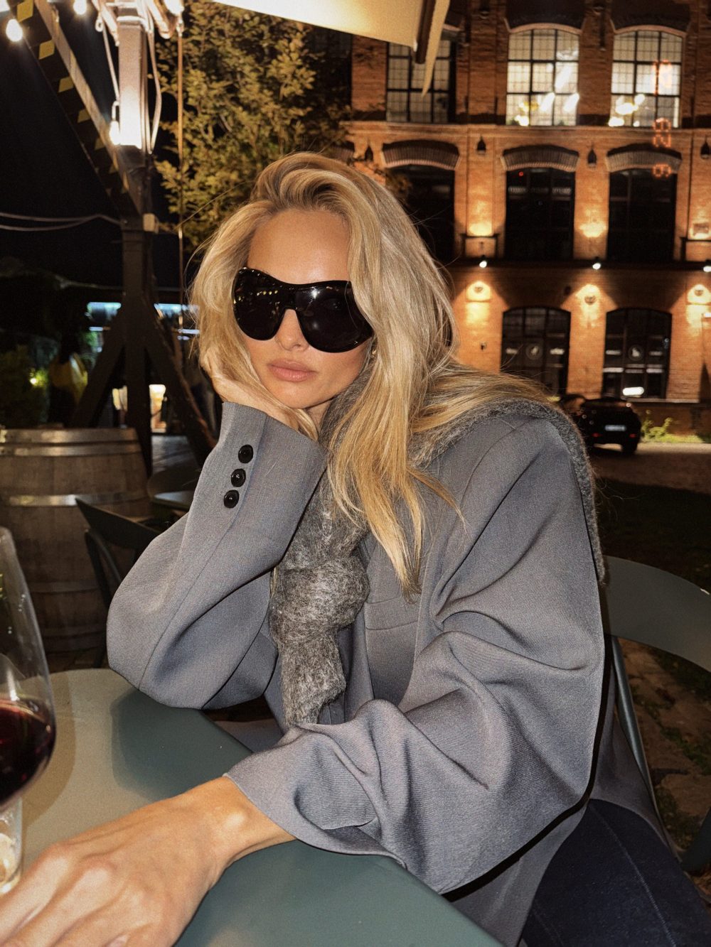 Woman with long blonde hair wearing oversized black sunglasses and a gray coat, resting her head on her hand at an outdoor cafe table at night; a brick building with warm lights in the background, and a glass of red wine on the table.