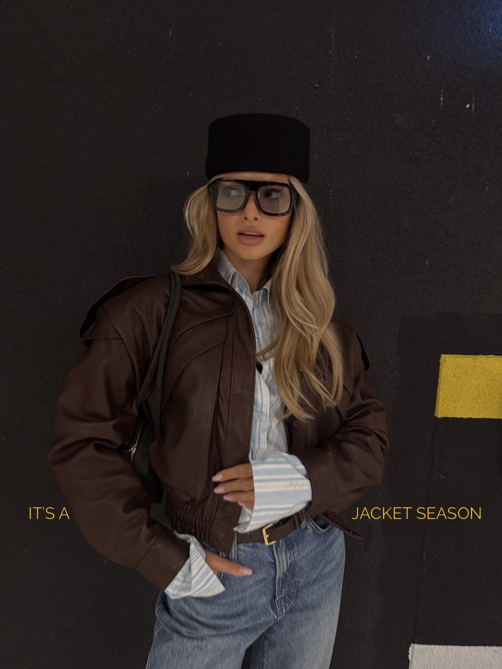 Fashion photo of a woman in a brown leather jacket, striped shirt, blue jeans, oversized sunglasses, and a tall black hat, posed against a dark wall with the caption 'It's a jacket season' visible at the bottom.