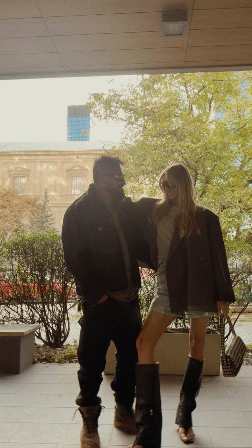 Two people posing together outdoors under a covered area, wearing jackets and boots with a leafy courtyard and building behind them.