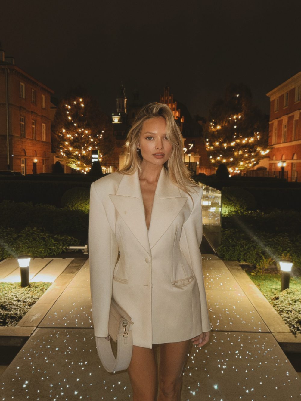 Woman posing in a cream blazer dress on a lit garden path at night, with string lights in the background.