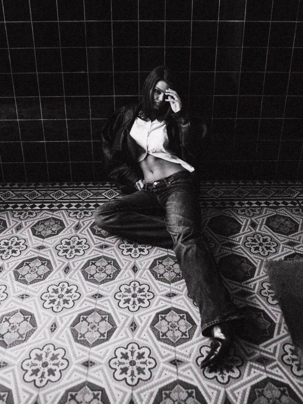 Black-and-white portrait of a woman sitting on patterned floor tiles, wearing a leather jacket and cropped white shirt, hand at her face.