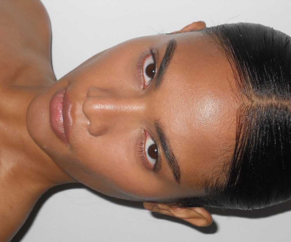 Close-up of a woman's face in profile, showing her eye, eyebrow, and smooth skin with dark hair pulled back against a light background.